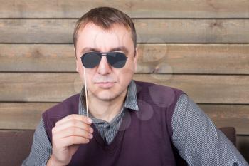 Playful man with funny sunglasses on a stick, wooden background. Fun photo props and accessories for photo shoots