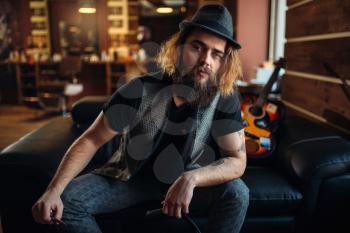 Bearded barber sitting on black leather couch, guitar and  barbershop inerior on background
