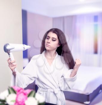 Beautiful woman in bathrobe drying her hair with hairdryer against mirror in bedroom