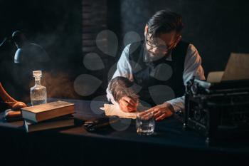 Bearded writer in glasses writes with a feather. Retro typewriter, crystal decanter, books and vintage lamp on the desk