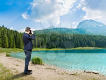 Male photographer taking picture of lake on digital camera, green wood and mountains on background