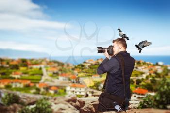 Male photographer with dove sitting on his head takes pucture on digital camera with professional lens standing on mountain