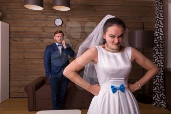 Portrait of dissatisfied bride against groom talking on the phone, wooden room on background.