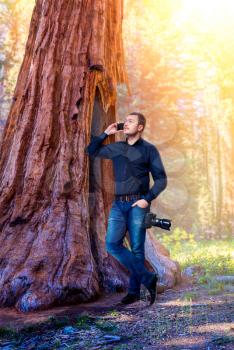 Photographer with professional digital camera talking by mobile phone leaning on the trunk of a large tree, forest on background
