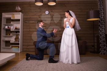 Groom in suit holding wedding rings, standing on his knees and making a proposal of marriage to happy bride in white dress and veil