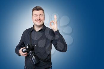 Professional photographer with digital camera shows ok sign, blue background