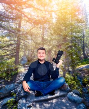 Smiling photographer with camera sitting in a yoga pose, pine forest on background