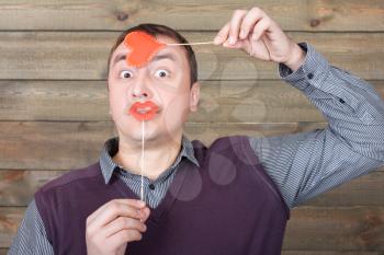 Young man with funny red lips and heart on a sticks in hand, wooden background. Fun photo props and accessories for shoots
