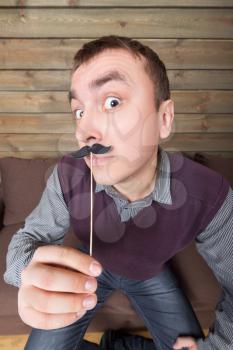 Young man with funny moustache on a stick, wooden background. Fun photo props and accessories for shoots