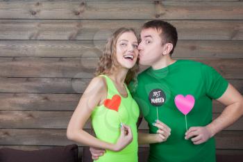Man kisses woman and shows funny icons on a sticks with hearts and just married inscription, wooden background. Fun photo props and accessories for shoots