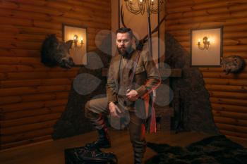 Respectable hunter man with old gun in retrro style traditional hunting clothing standing against antique chest. Retro rifle, vintage clothes. Room with fireplace, stuffed wild animals, bear skin and 