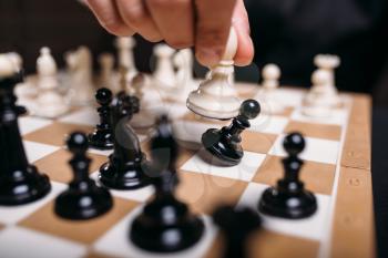 Male chess player hand holding white figure, closeup. Chessplayer makes a move and eats black pawn, selective focus. Intelligence battle on chessboard concept