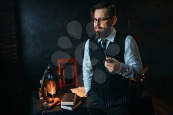 Serious bearded writer in glasses smoking a pipe. Feather, crystal decanter, books and vintage lamp on the desk
