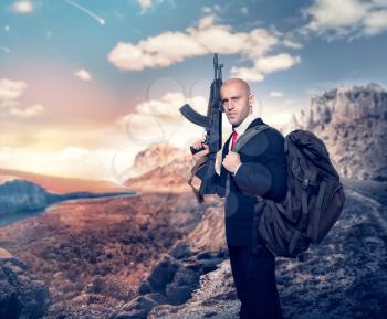 Professional agent on secret mission concept. Hired murderer in suit and red tie with backpack holding machine gun in hand, desert valley on background
