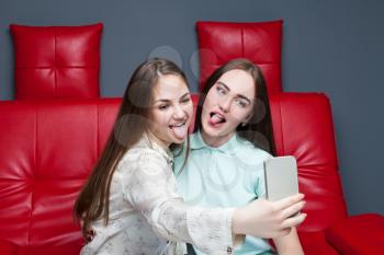 Two smiling women sitting on red leather couch and makes selfie on phone camera. Female friendship. Leisure of happy girls 