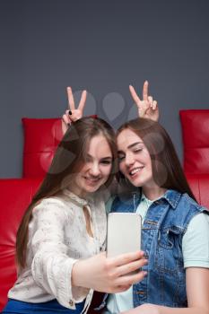 Two funny girlfriends sitting on red leather couch and makes selfie on phone camera. Female friendship. Leisure of happy girls 