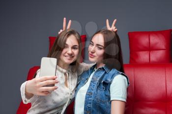 Two funny girlfriends sitting on red leather couch and makes selfie on phone camera. Female friendship. Leisure of happy girls