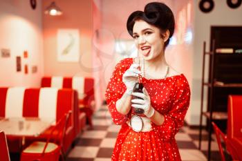 Pinup girl with make-up drinking popular carbonated drink in retro cafe, 50 american fashion. Red dress with polka dots, vintage style