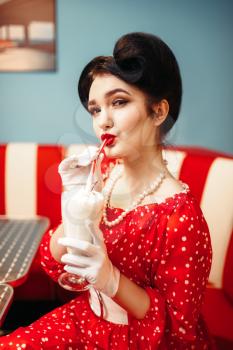 Sexy pin up girl with make-up drinks milkshake through a straw in retro cafe, popular american fashion 50s and 60s. Red dress with polka dots, vintage style