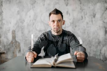 Strange freak man with fork and knife in hands eats the sheets from the book. Alone guy in abandoned house, grunge room interior. Mad male person, crazy guy