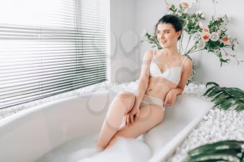 Attractive woman in white underwear sitting in bath with foam. Bathroom interior with window on background