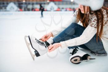 Young woman sitting on ice and ties the shoelaces on skates, skating rink. Winter ice-skating on open air, active leisure