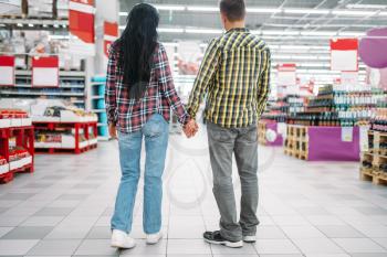 Young couple hold hands in supermarket, back view. Male and female customers on family shopping. Man and woman purchasing in hypermarket