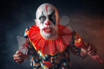Mad bloody clown with meat cleaver and human hand. Man with makeup in halloween costume, crazy maniac, horror