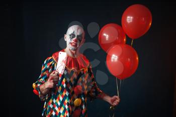 Ugly bloody clown with meat cleaver holds air balloon, horror. Man with makeup in carnival costume, crazy maniac