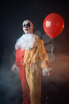Mad bloody clown with makeup in carnival costume holds air balloon, crazy maniac, scary monster