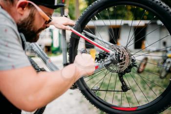 Bicycle mechanic in apron adjusts with service tools back disk brakes. Cycle workshop outdoor. Bicycling sport, bearded repairman