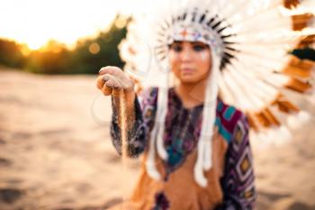 Beautiful woman in a suit of the American Indian play with sand on sunset. Young girl in headdress made of feathers of wild birds