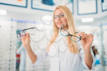Female optician standing against showcase with glasses in optics store. Selection of eyeglasses with professional optometrist
