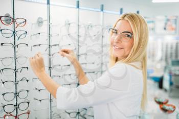 Female optometrist shows glasses in optics store. Selection of eyeglasses with professional optician, optometry