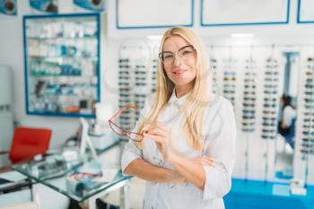 Female optometrist holds spectacles in hands, showcase with glasses in optics shop on background. Selection of eyeglasses with professional optician
