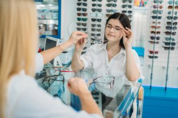 Female optician and consumer chooses glasses frame in optics store. Selection of spectacles with professional optometrist