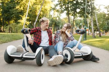 Young couple sitting on road with gyro board in summer park. Outdoor recreation with electric gyroboard. Eco transport, electrical gyroscope vehicle