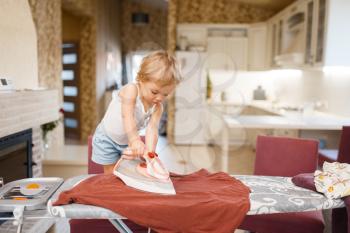 Little baby irons clothes on the kitchen. Kid doing housework at home. Young housewife clean the house