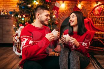 Love couple, romantic christmas celebration. Xmas holidays, man and woman sitting on the floor and drinks coffee with whipped cream, festive decoration on background
