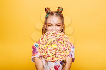 Beautiful young woman with huge candy instead of a head. Big colorful caramel lollypop in hands of woman. Portrait of attractive lady with big lollypop, yellow wall on background.