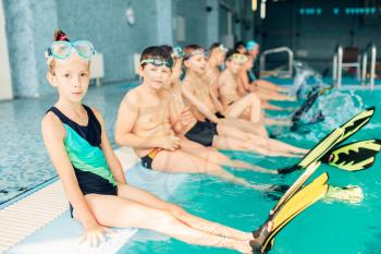 Portrait of children with colorful flippers sits near pool. Boys and girls ready to swim in pool. Happy kids in modern sport center. Concept of fun, leisure and recreation.