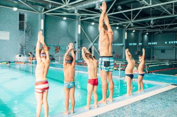 Instructor with children stands near water in pool and preparing for diving. Trainer shows kids exercise, view from back. Modern sports center on background. Sportive kids activity.