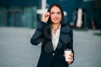 Portrait of young, beautiful business woman in glasses and suit with laptop in hands. Modern building, financial center, cityscape. Successful female businessperson