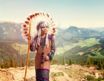 American Indian girl in traditional costume against mountain valley, Cherokee, Navajo. Headdress made of feathers of wild birds