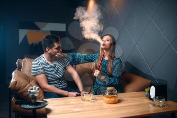Young woman smokes hookah against her man at the bar, tobacco smoking and night relaxation of love couple