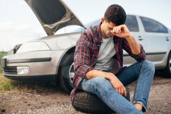 Tired man sitting on tire, broken car with open hood on background
