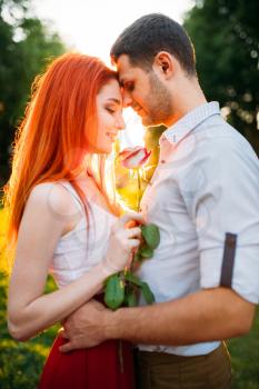 Beautiful love couple hugs in summer park on sunset. Attracrive woman with rose and young man leisure together outdoors. Romantic date