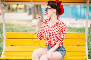 Sexy pinup girl sitting on bench in park and licks sweet lollipop, retro american fashion. Attractive model in pin up style