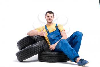 Serviceman in blue uniform sitting on heap of tires, white background, repairman with tyres