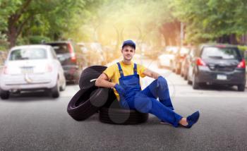 Serviceman in uniform sitting on a pile of tires, street road on background. Repairman, wheel mounting, tyre service